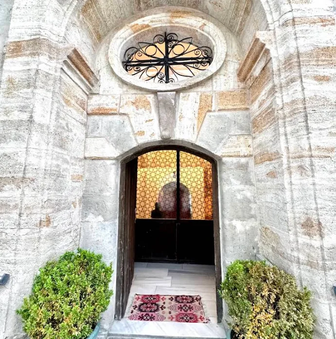 Historic cut-stone entrance of Ürgüp Hamam with an oval ironwork window and traditional rug.
